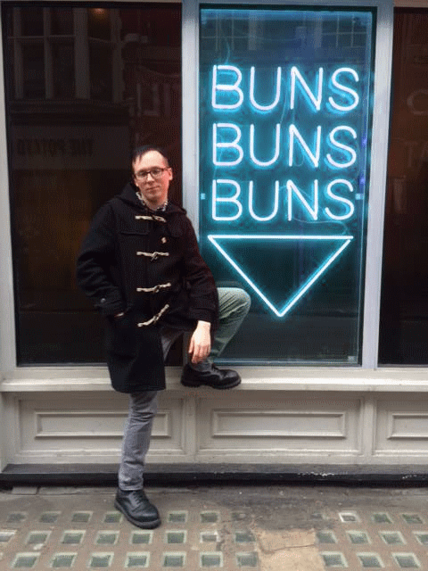The author wears a duffle coat, chinos, and doc martens, standing casually before a window with a flashing neon sign that reads BUNS BUNS BUNS.